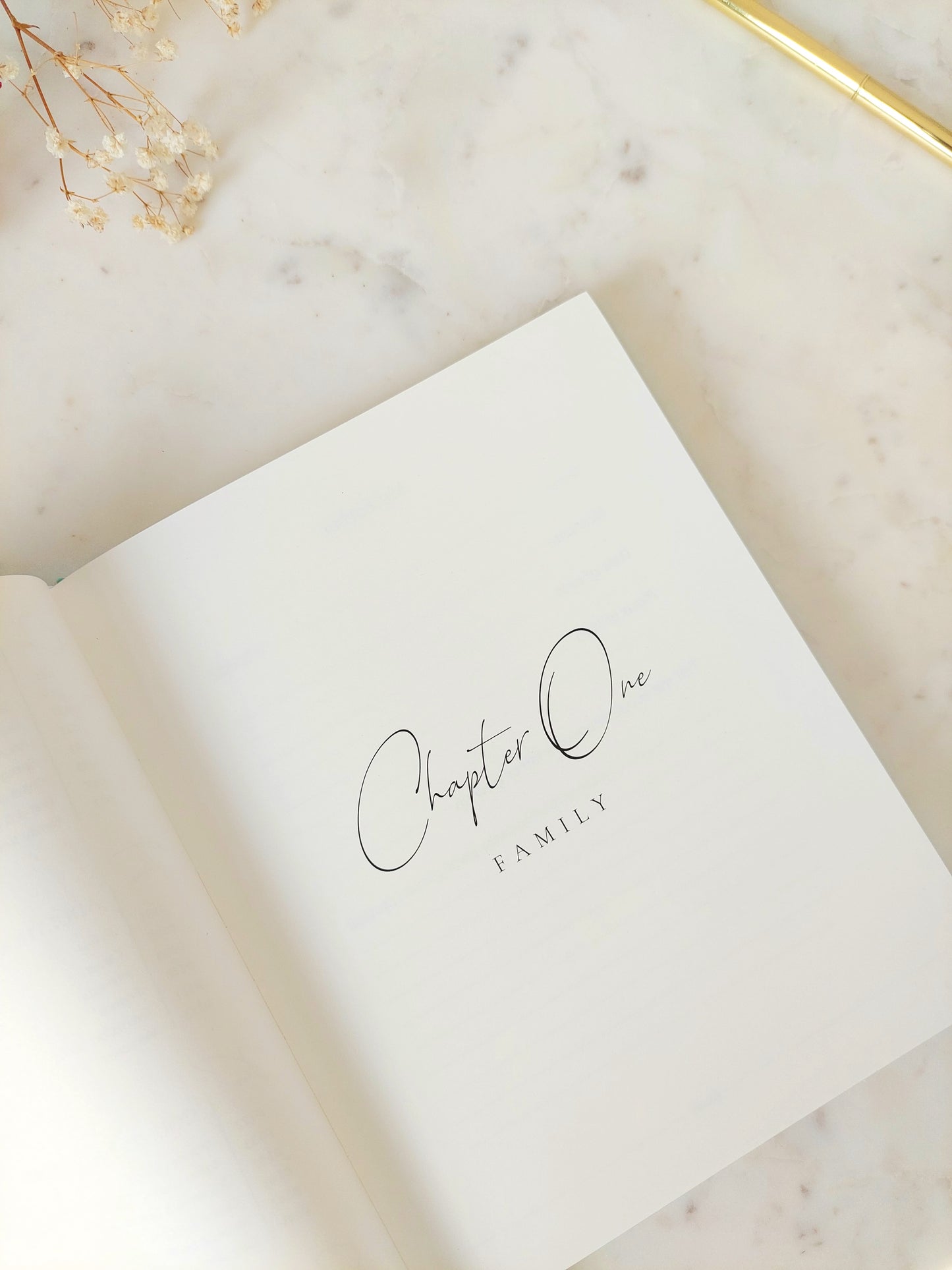 Inside the legacy journal showing the title page for the first chapter. Black font on subtle ivory paper. Chapter One is written in easy to read calligraphy. Underneath it reads FAMILY. The book is on a neutral white marble table with a gold pen