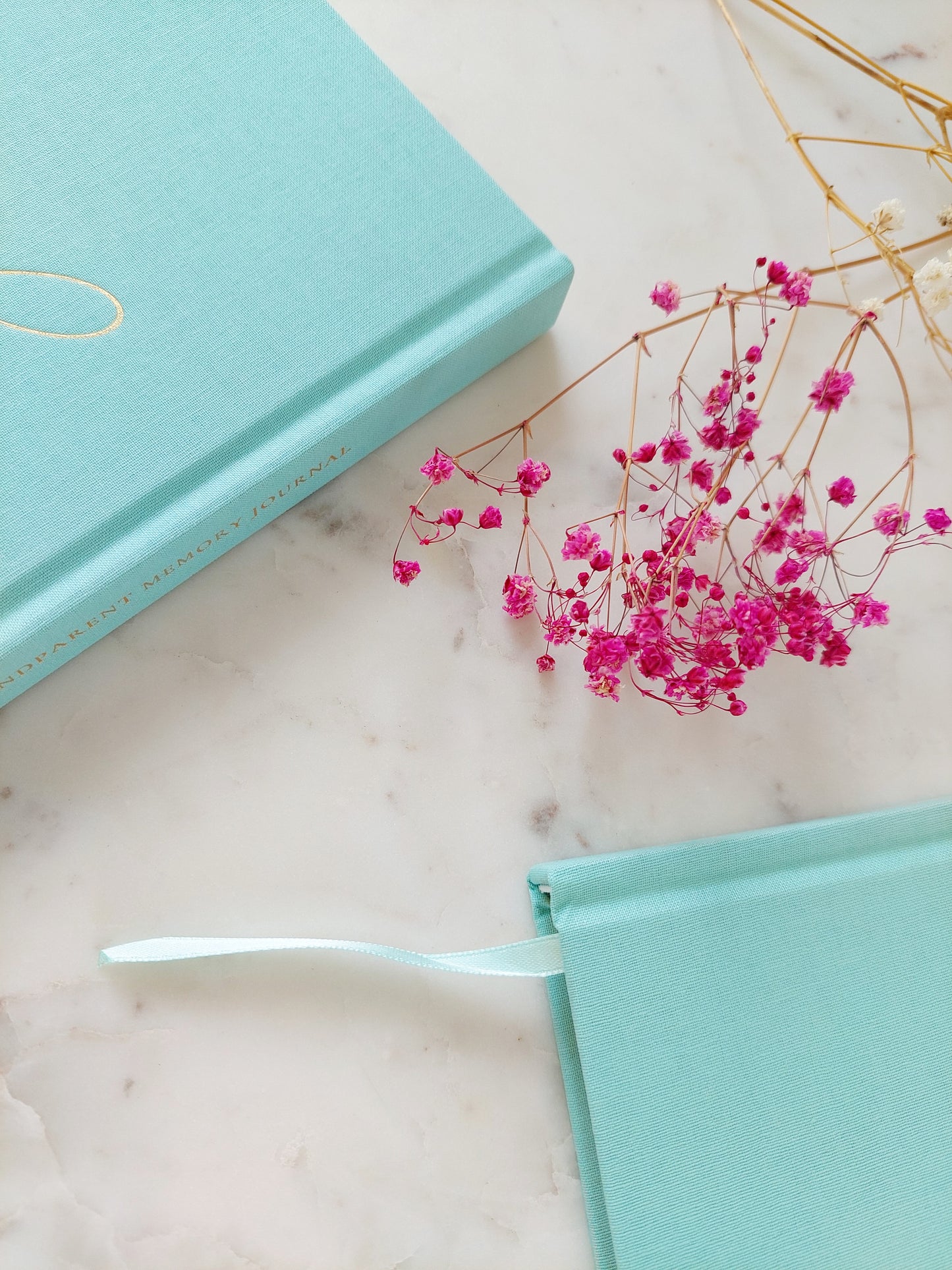 A close up of two aquamarine journals next to each other on a white marble table from a bird’s eye view. Shows that the book has an aquamarine ribbon bookmark