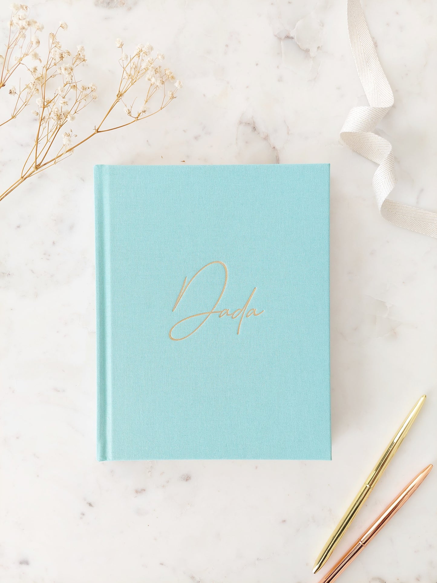 Birds eye view of the front of a journal; its colour is aquamarine. It is a hardback linen book with Dada written in gold foil, in easy to read calligraphy style writing. On a neutral white marble table with two gold pens beside the book