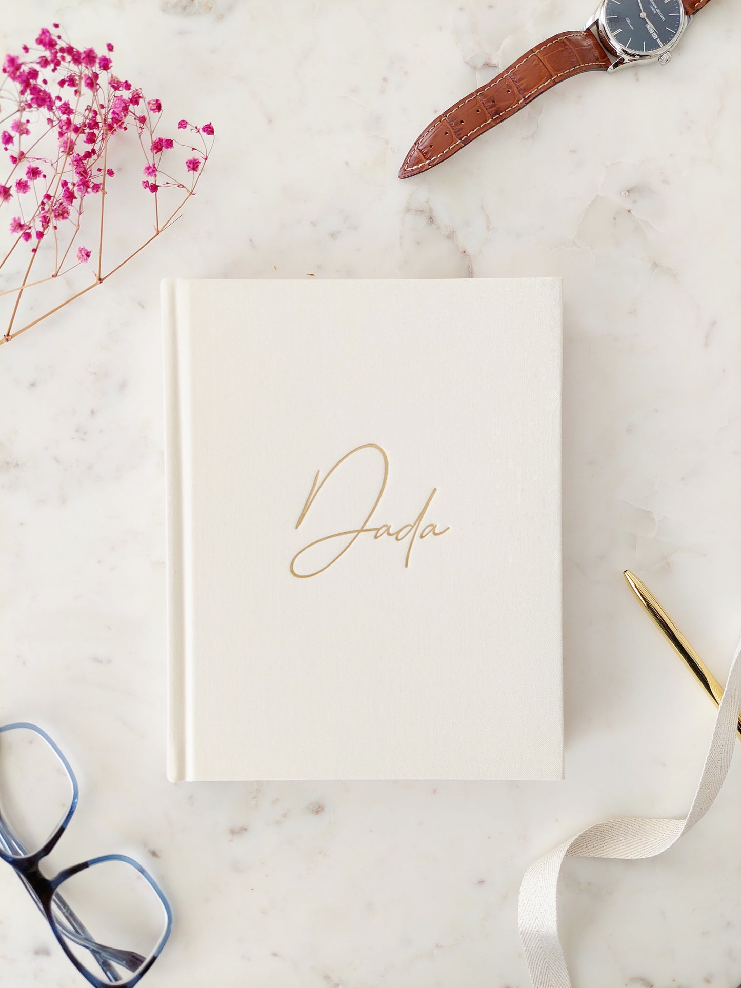 Bird’s eye view of the front of a journal. It is a hardback, ivory linen book with Dada written in gold foil, in easy to read calligraphy style writing. On a neutral white marble table with a gold pen & spectacles beside the book showing the scale