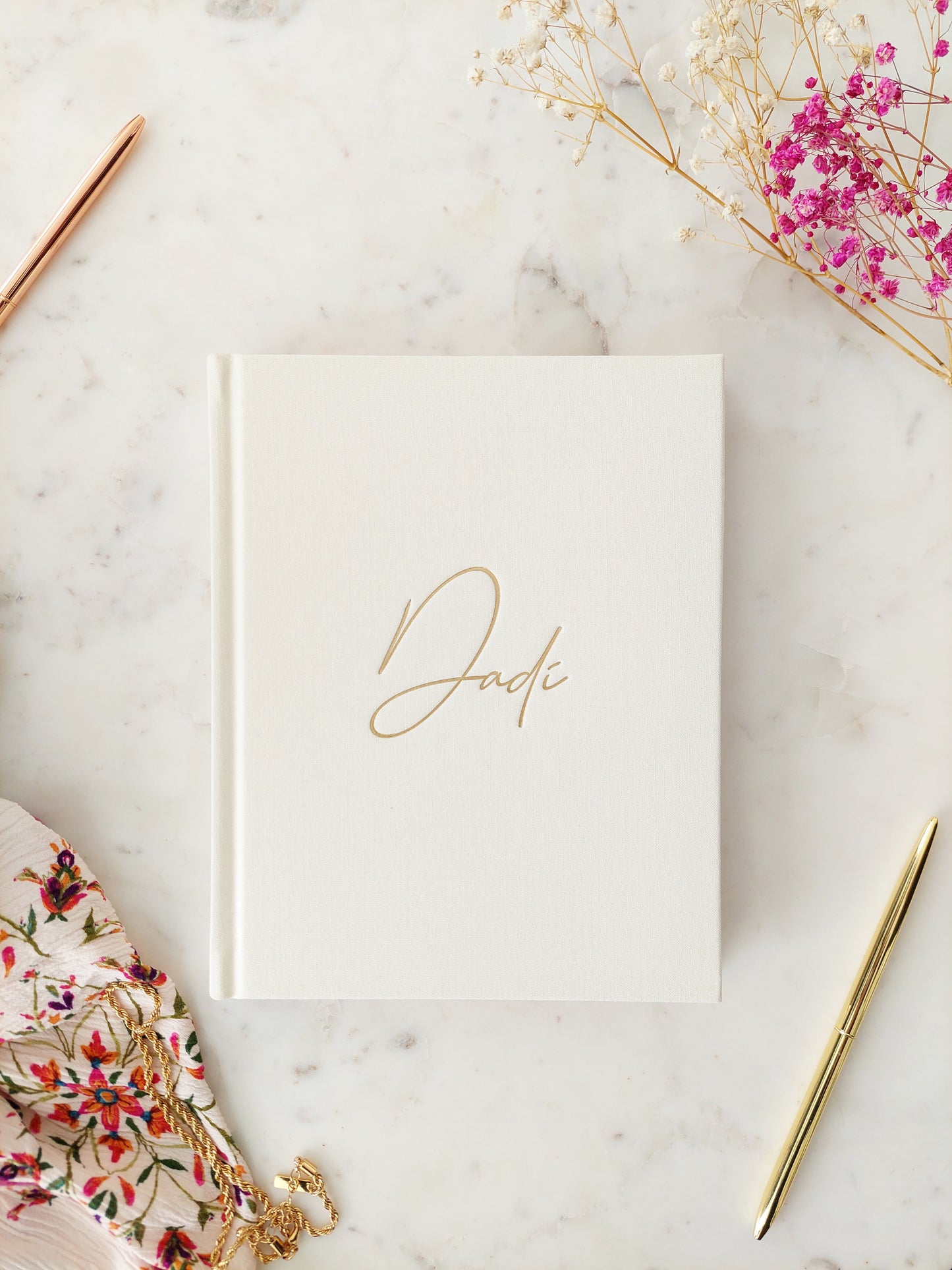 Bird’s eye view of the front of a journal. It is a hardback, ivory linen book with Dadi written in gold foil, in easy to read calligraphy style writing. On a neutral white marble table with a gold pen, necklace and floral scarf beside it