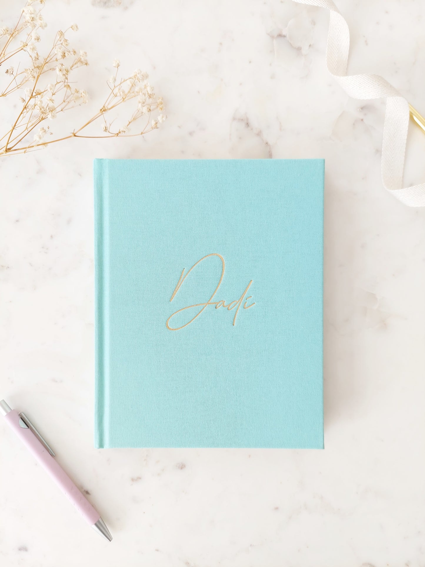 Bird’s eye view of the front of a journal; its colour is aquamarine. It is a hardback linen book with Dadi written in gold foil, in easy to read calligraphy style writing. On a neutral white marble table with a pink pen and white ribbon beside it