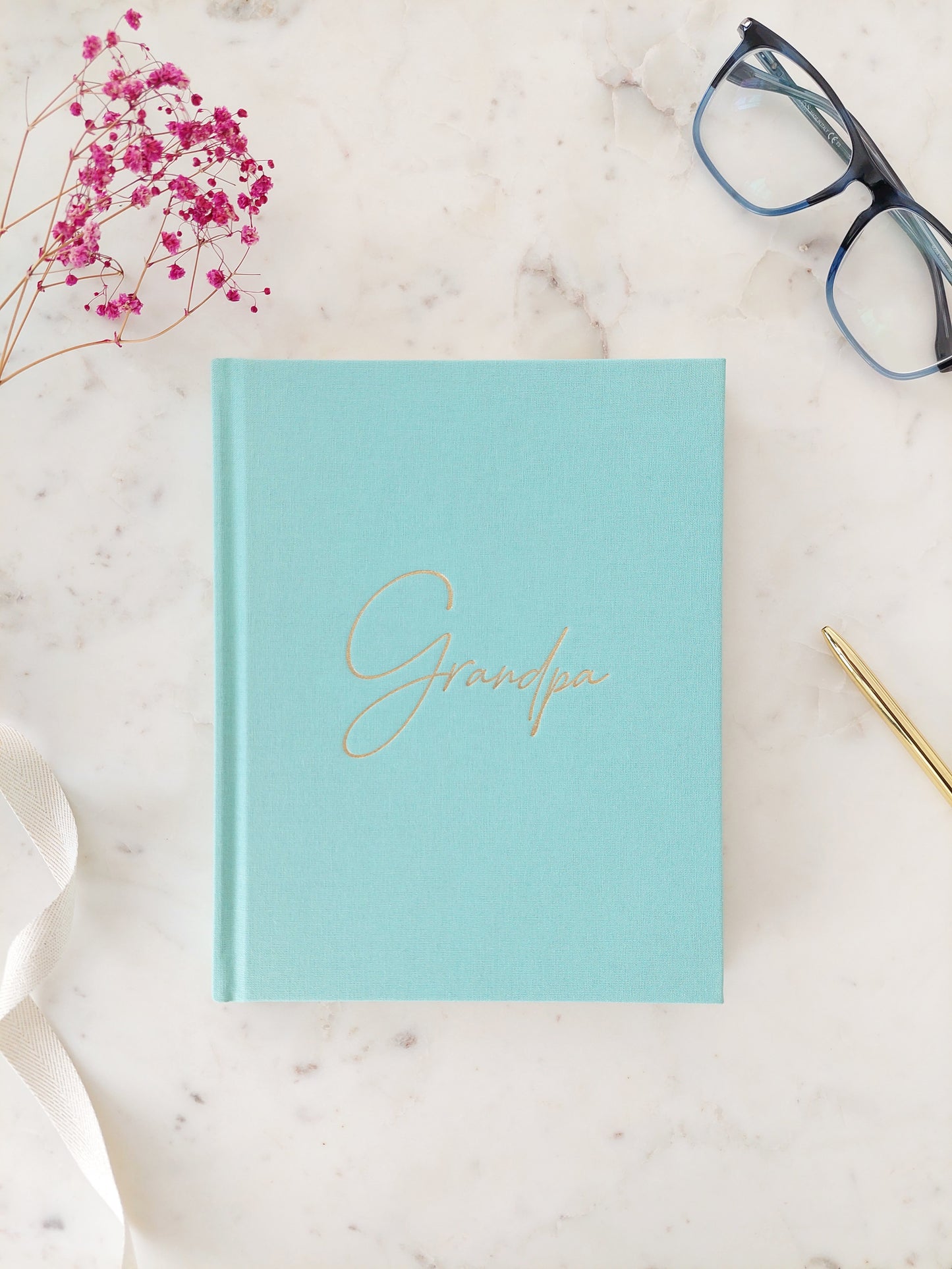 The front of a journal; the colour is aquamarine. It is a hardback linen book with Grandpa written in gold foil, in easy to read calligraphy style writing. On a neutral white marble table with a gold pen & spectacles beside the book showing the scale