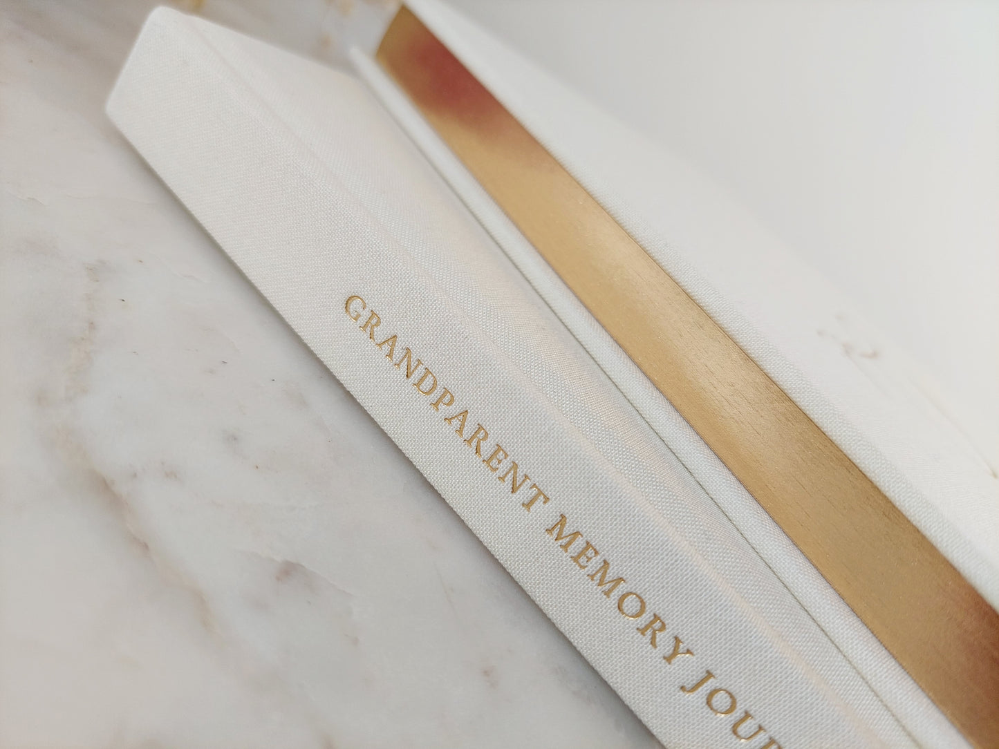 A close up of two ivory journals (one on top of the other) from the side view. One shows the text Grandparent Memory Journal in gold foil on the spine. The other shows the gold gilded edges on the paper edges. The linen texture can be seen.