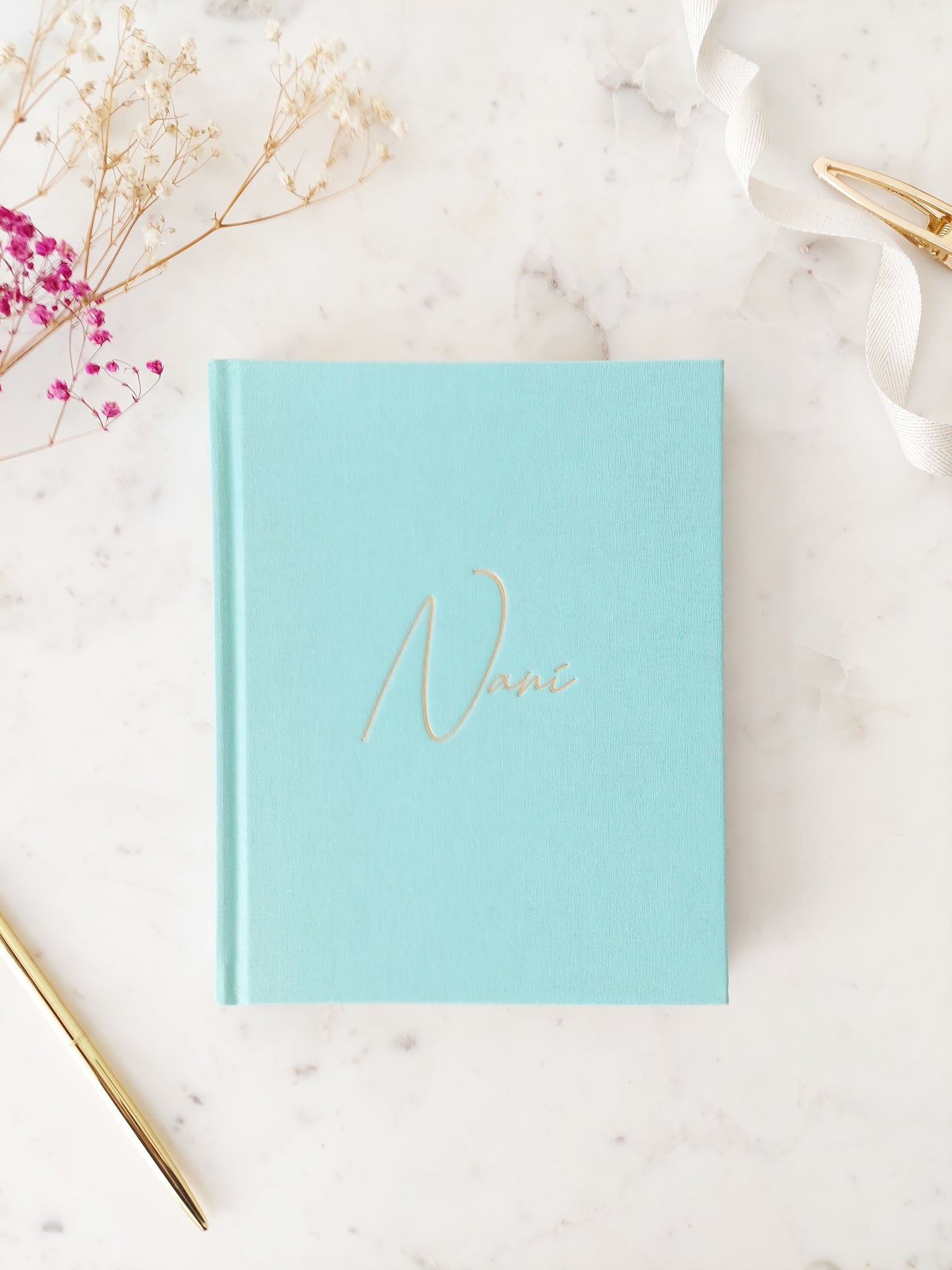 Bird’s eye view of the front of a journal; its colour is aquamarine. It is a hardback linen book with Nani written in gold foil, in easy to read calligraphy style writing. On a neutral white marble table with a gold pen and white ribbon beside it.