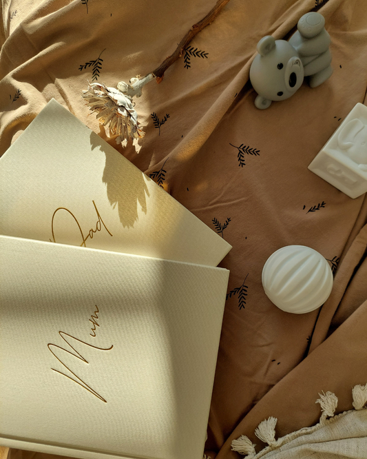 Two ivory linen life story journals with 'Mum' 'Dad' written in gold foil cursive font. Displayed on a woodland brown baby blanket