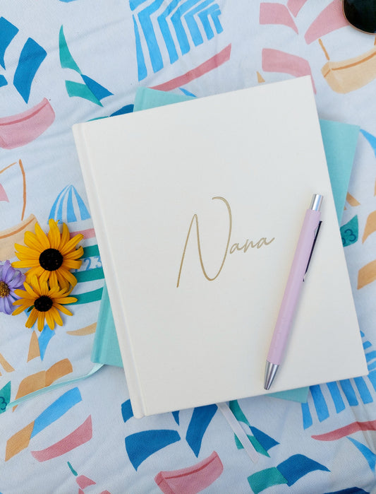 An ivory hardback grandparent life story journal with 'Nana' written in gold. Displayed on a colourful patterned background with sunflowers 