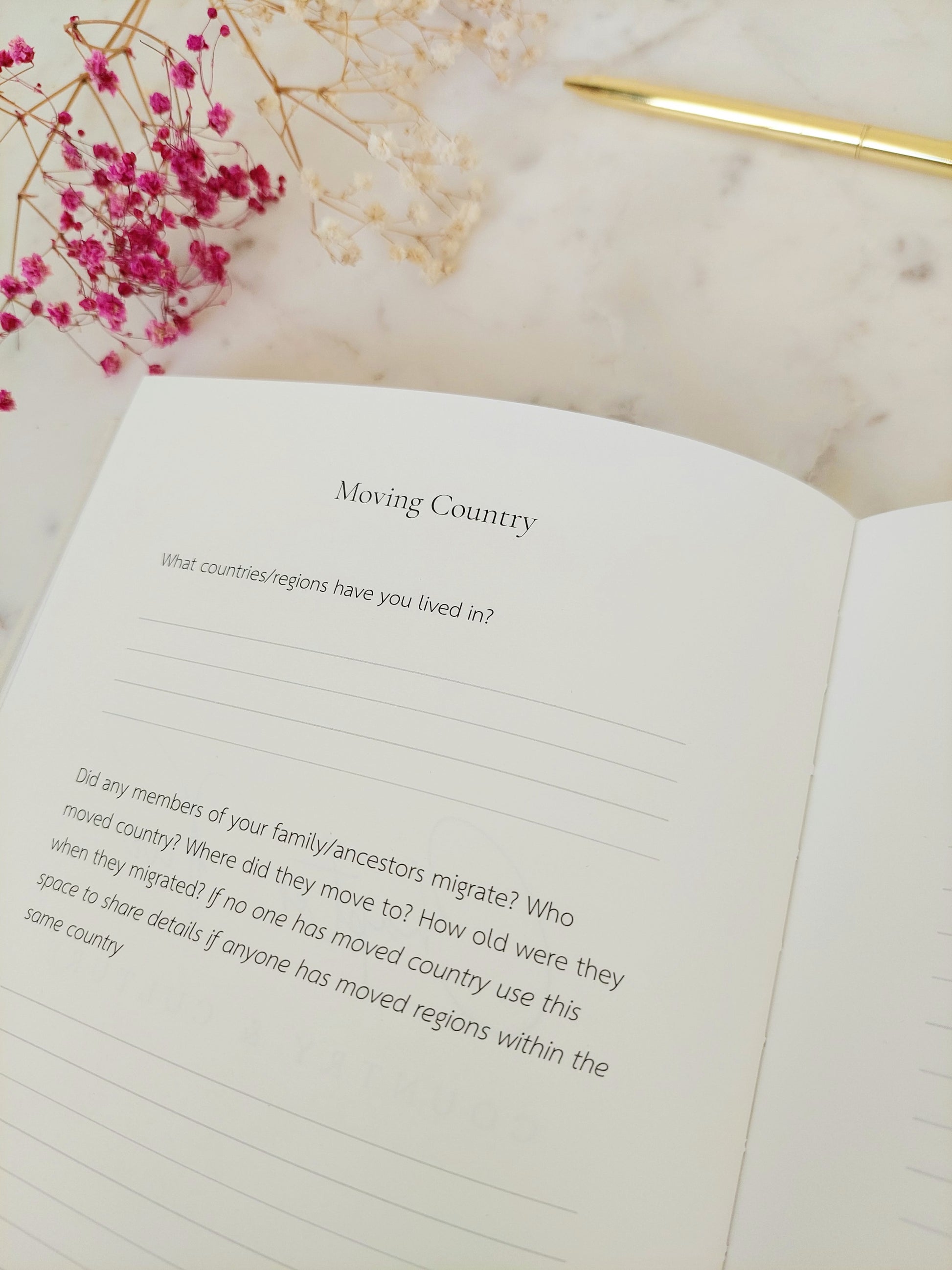 The life story book is open on a white marble table, there is a gold pen and gypsophila. The page heading reads Moving Country followed by two questions with lines underneath each to complete the answers. Font is black on subtle ivory paper.