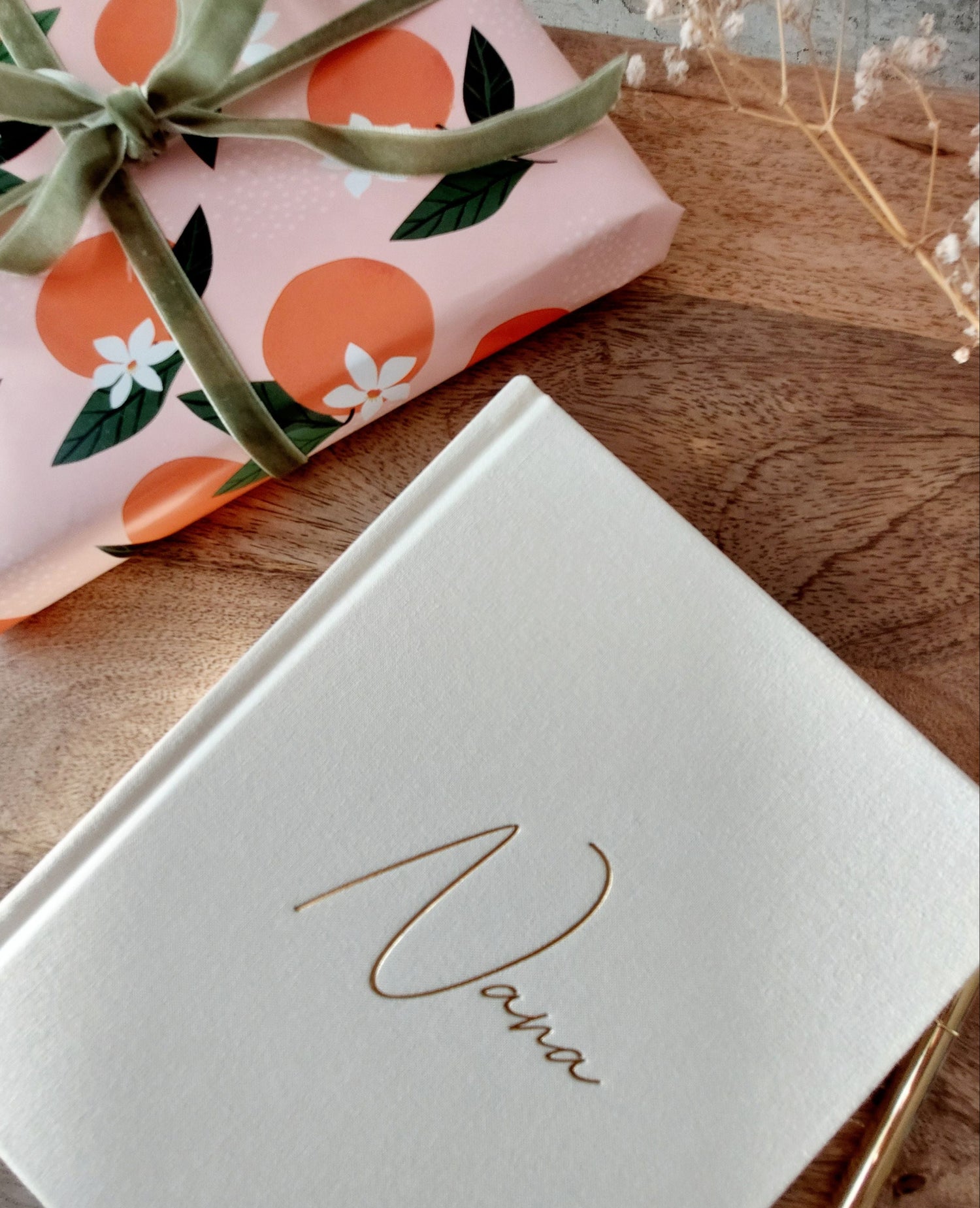 Ivory journal with 'Nana' written on front on a wooden surface next to a pink and orange floral-patterned wrapped book with green ribbon.