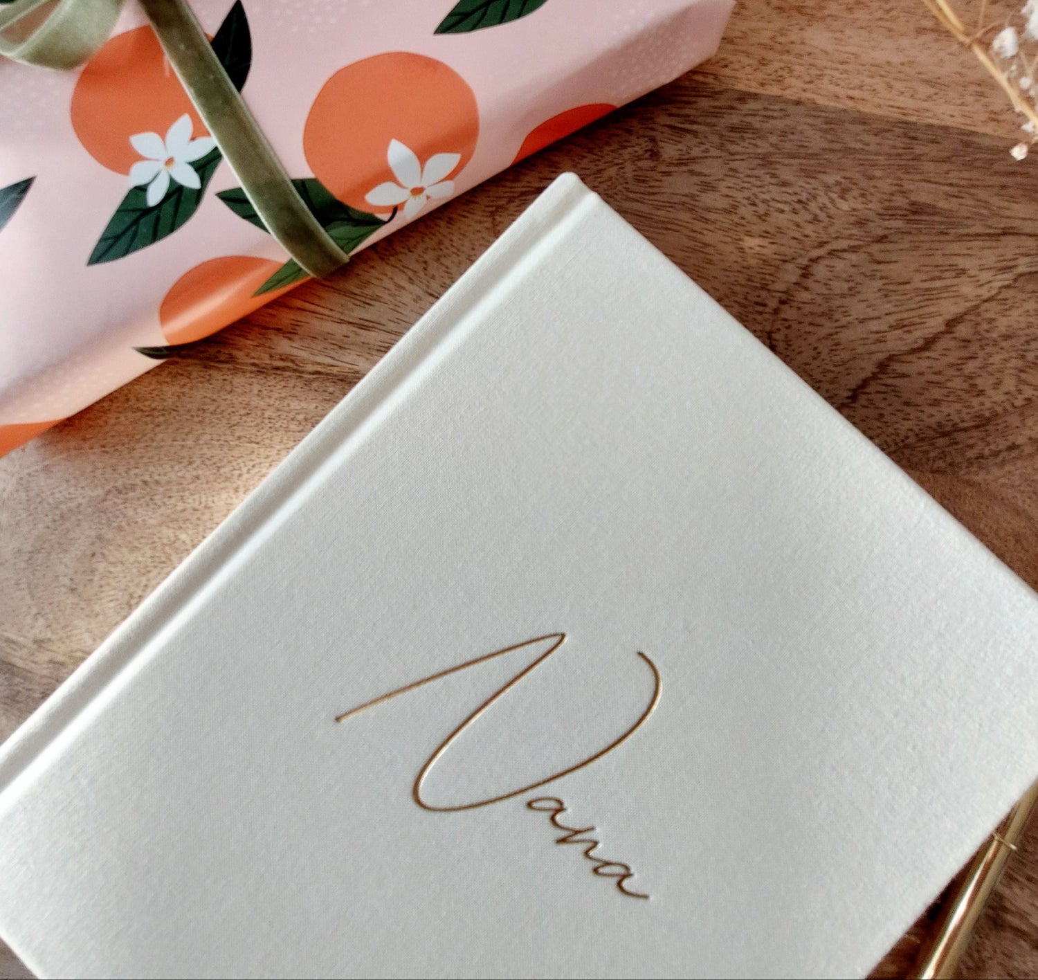 Linen journal with 'Nana' in gold cursive font, on a wooden surface next to a pink gift box with orange pattern and green ribbon.