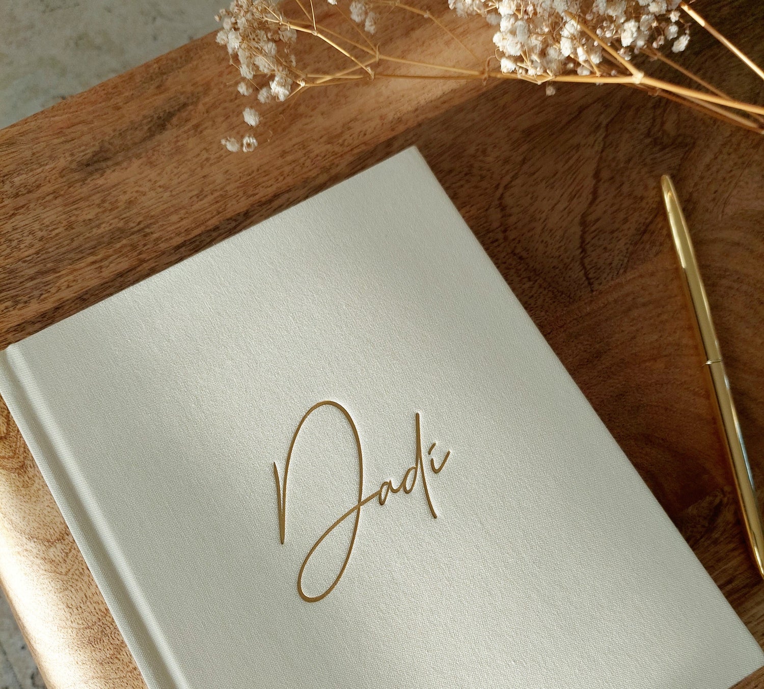 Ivory linen book with 'Dadi' written in gold on a wooden surface with dried flowers.