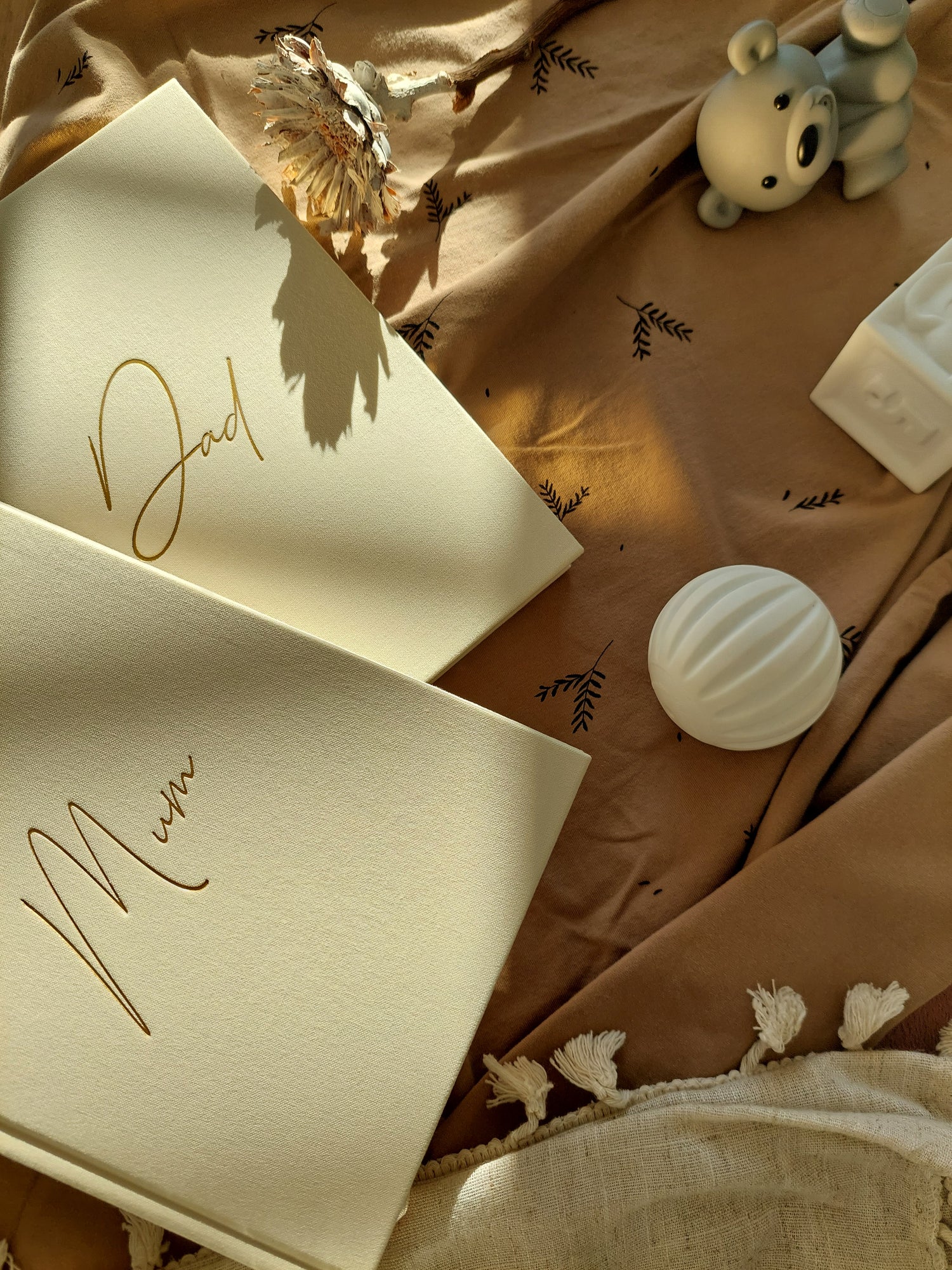 Two ivory legacy journals with gold text 'Mum' and Dad' on a textured surface with decorative baby items.