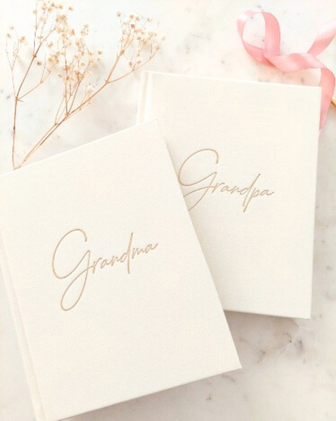 Two ivory journals with 'Grandma' and 'Grandpa' text in gold on a marble surface with dried flowers and a pink ribbon.